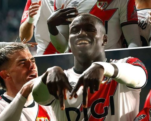 Nobel Mendy celebrates a goal with teammates, smiling and making a hand gesture while wearing the Rayo Vallecano home kit with DIGI sponsor.