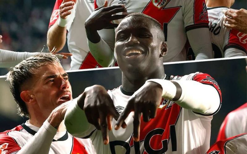 Nobel Mendy celebrates a goal with teammates, smiling and making a hand gesture while wearing the Rayo Vallecano home kit with DIGI sponsor.